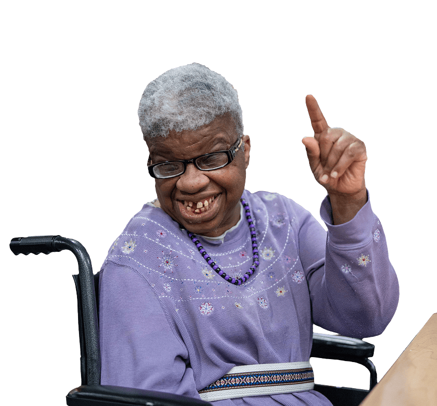 Woman in a purple sweater and grey hair in a wheel chair, smiling at the camera with her pointer finger in the air