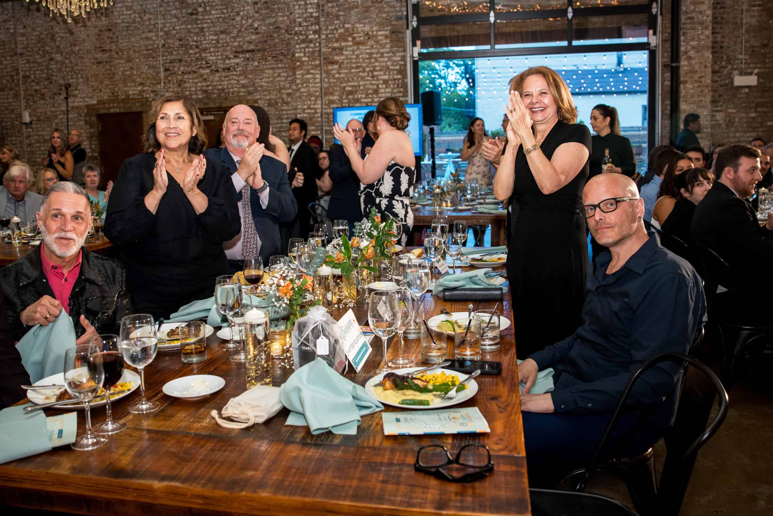 A group of people gathered around a table, clapping enthusiastically, at a dinner party for the Trailblazers event