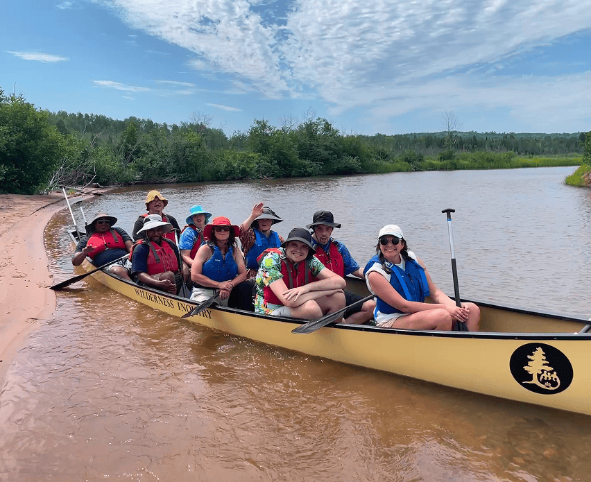 Nine people in a canoe, all wearing hats and smiling at the camera